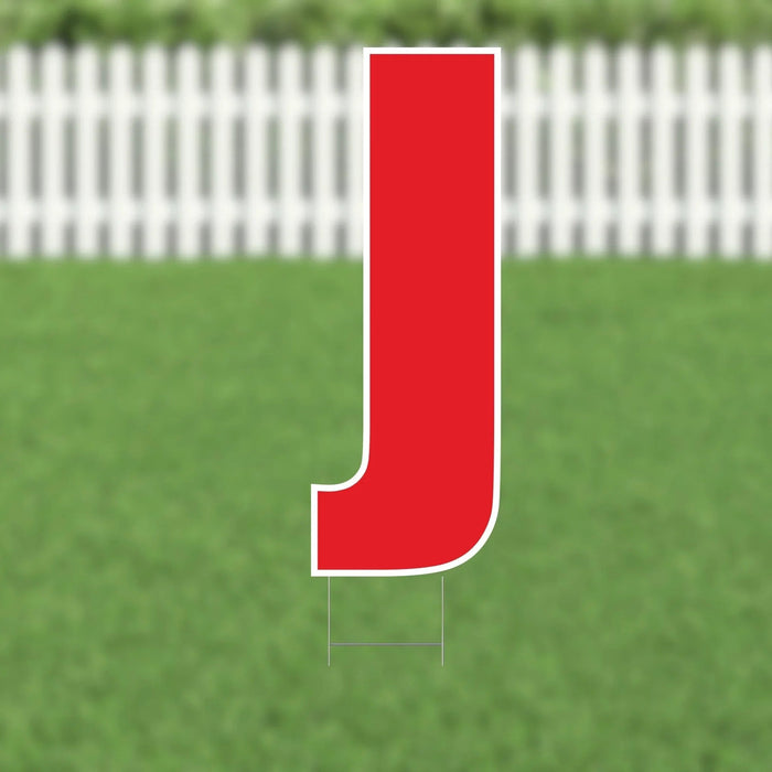 Red letter J yard sign with white border and metal stakes, displayed on a lawn with white picket fence in background.