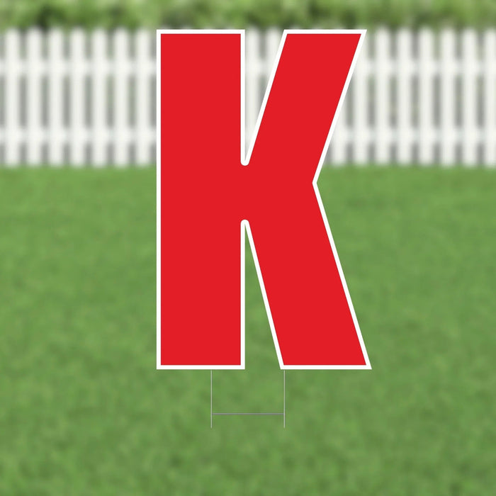 Large red letter K with white outline and metal stakes, displayed on a lawn with white picket fence in background.
