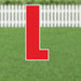 Red letter L yard sign with white border and metal stakes, displayed on green grass with white picket fence background.