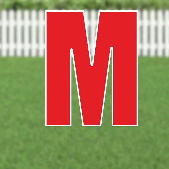 Large red letter M yard sign with white border and metal stakes, displayed on a grassy lawn with white picket fence in background.