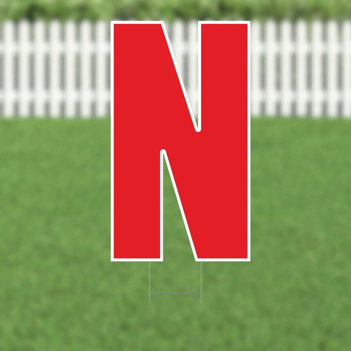 Large red letter N yard sign with white border and metal stakes, displayed on green grass with white picket fence background.