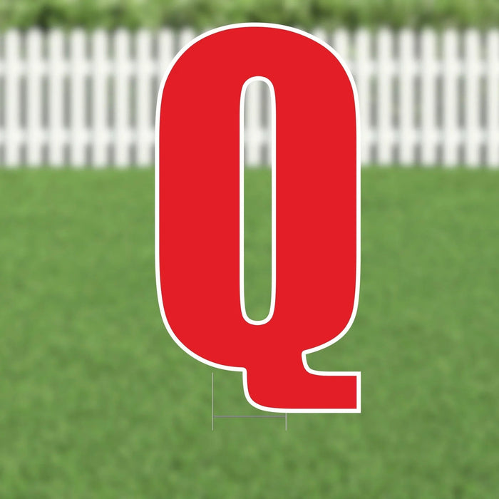 Red letter Q yard sign with white border and mounting stakes, displayed on grass with white fence background.