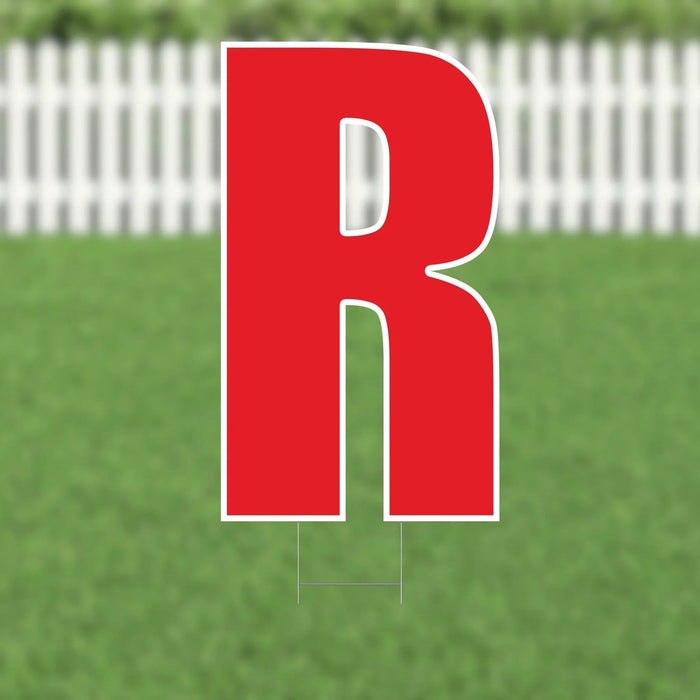 Large red letter R yard sign with white outline and metal stakes, displayed on green grass with white picket fence background.