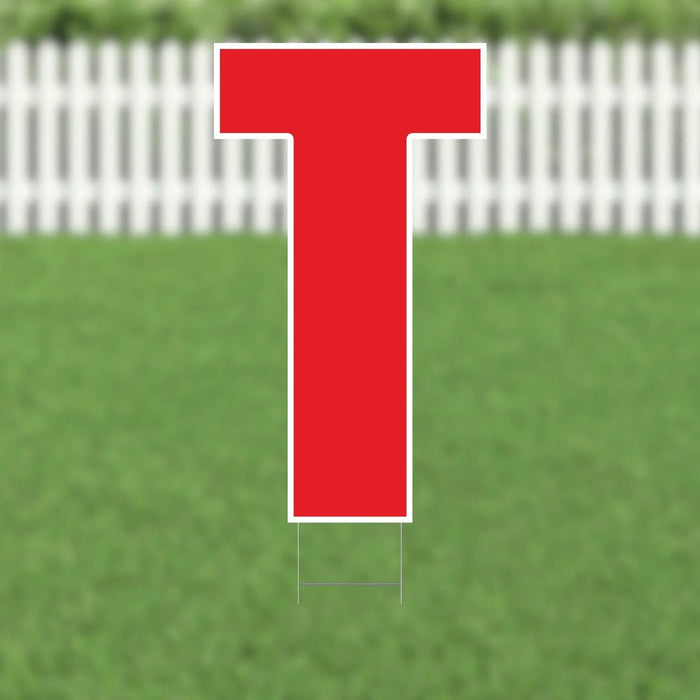 Red letter T yard sign with white border and metal stakes, displayed on green grass with white picket fence background.
