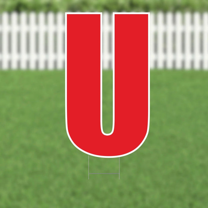 Red capital letter U yard sign with white border and metal stakes, displayed on green grass with white picket fence background.
