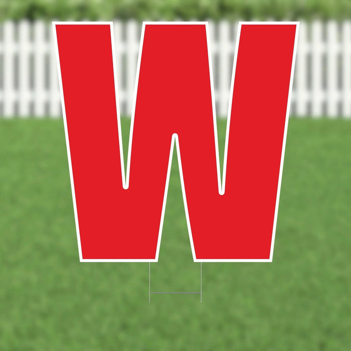 Red letter W yard sign with white border and metal stakes, displayed on grass with white picket fence in background.