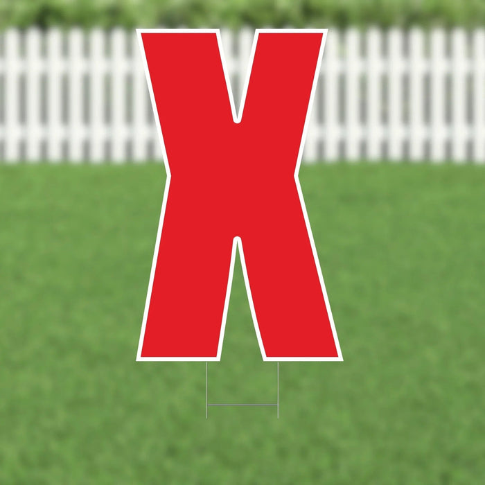 Large red letter X yard sign with white border and stakes, displayed on a lawn with white picket fence background.