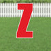 Red yard sign displaying the letter Z in large bold font, mounted on a green lawn with white picket fence in the background.