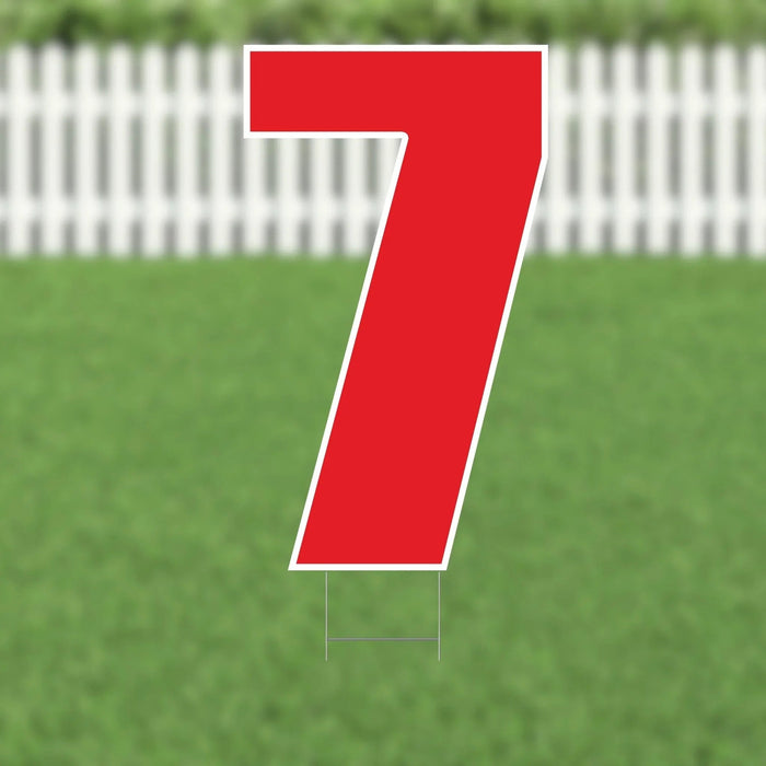 Large red number 7 yard sign with white border and metal stakes, displayed on a lawn with white picket fence in background.