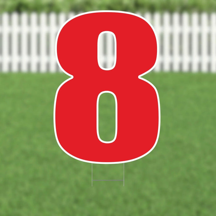 Large red number 8 yard sign with white outline and hollow centers, displayed on green grass with white picket fence in background.