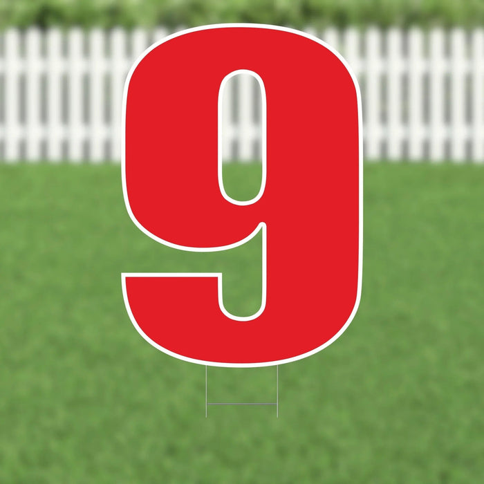 Large red number 9 yard sign with white outline and metal stakes, displayed on a lawn with white picket fence in the background.