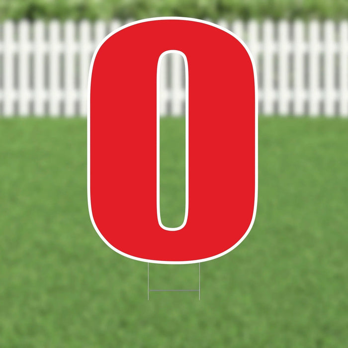Red number zero yard sign with white outline and metal stakes, displayed on grass with white picket fence in background.