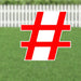 Red and white hashtag symbol yard sign with white border standing in grass with white picket fence in background.