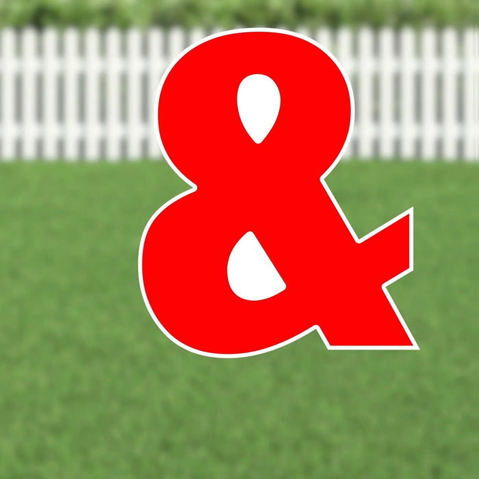 Large red ampersand symbol with white outline and cutout details, displayed in a yard against a white picket fence.