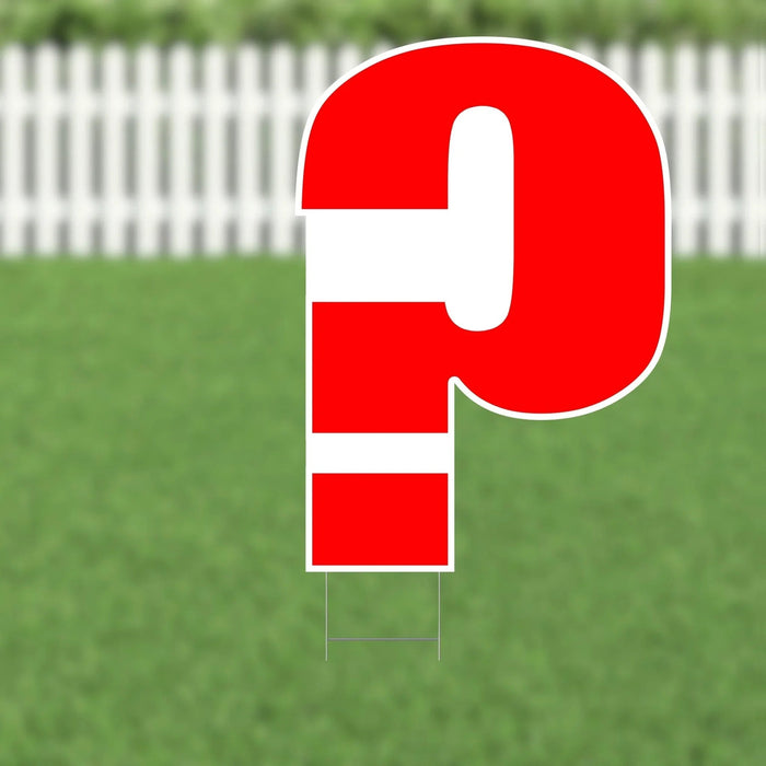 Red and white striped question mark yard sign with metal stakes, displayed on grass with a white picket fence in the background.